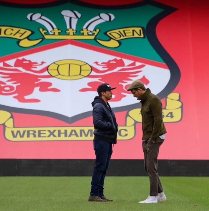 Two men talking to each other on a football pitch with the Wrexham AFC logo visible in the background