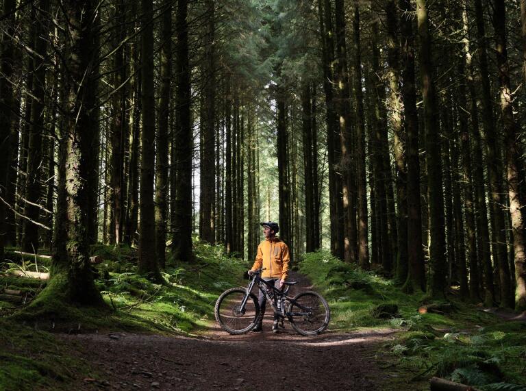 A person in a yellow jacket and biking gear standing with a mountain bike in a pathway surrounded by tall trees in a beautiful forest setting