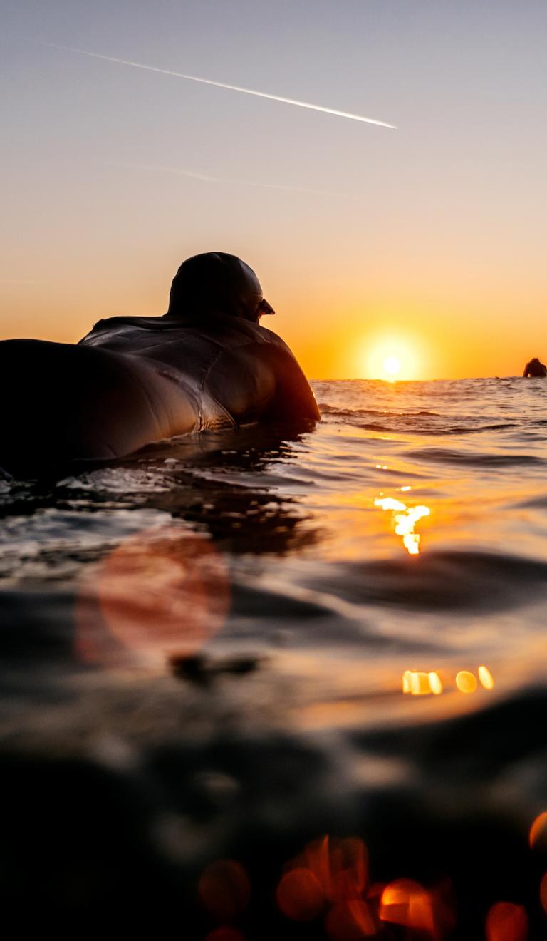 日が沈む中、サーフボードに横たわって波に乗るのを待っている人