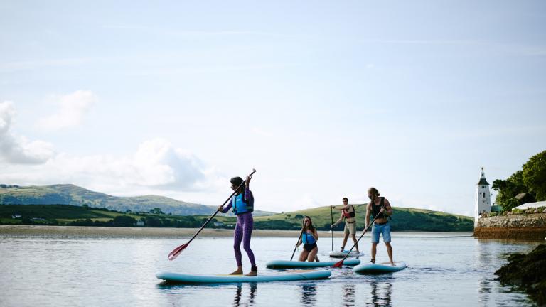 Des jeunes debout sur des paddles sur l'eau, entourés de douces collines et d'une tour.