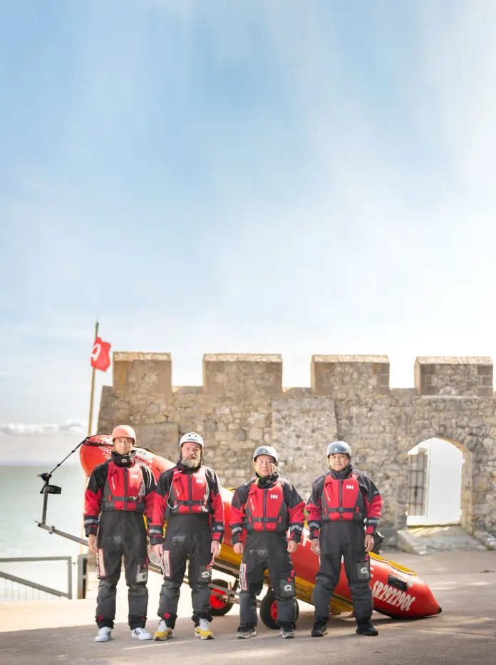 Four men dressed in sea rescue clothing stand in front of a stationary lifeboat on land in front of a castle wall