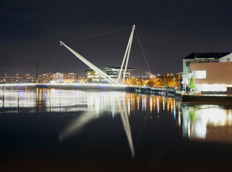 Foto nocturna de la pasarela con luces del edificio que se refleja en el río.