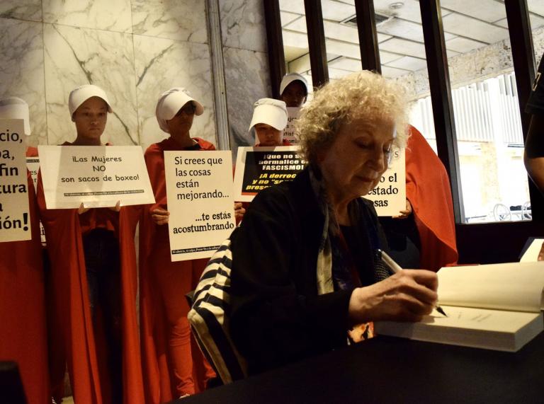 Margaret Atwood signiert Bücher, im Hintergrund Frauen in roten Umhängen.