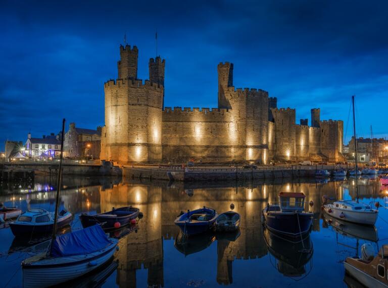 Eine große Burg mit Wasser und Booten im Vordergrund, beleuchtet gegen einen Nachthimmel.