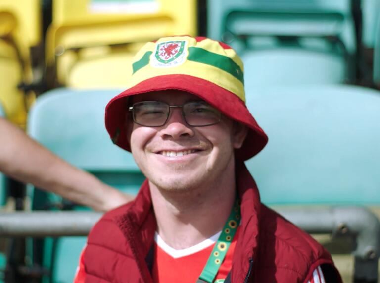 Un hombre sonriendo a la cámara con su camiseta y gorra de fútbol galés.