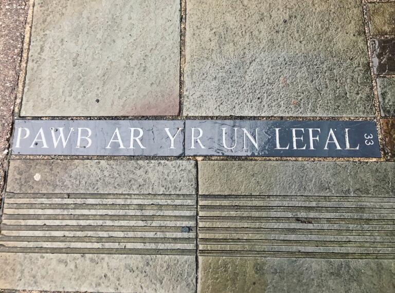 'Pawb ar yr un Lefel', escrito en un trozo de pizarra incrustado en un pavimento de losas.