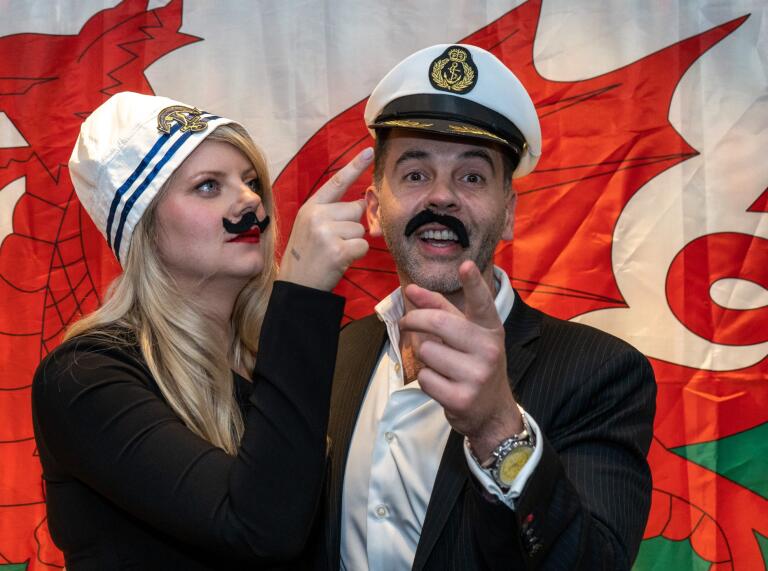Two people wearing sailor hats and fake moustaches standing in front of a Welsh flag
