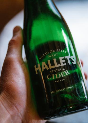 Una mano que sostiene una botella verde con Hallets Cider impresa.