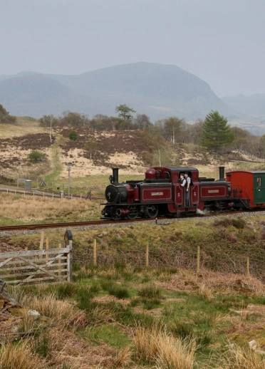 Une locomotive à vapeur rouge et verte roule le long d'une voie ferrée devant une toile de fond montagneuse.