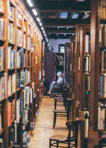 Una señora sentada y leyendo en una biblioteca, rodeada de estantes altos llenos de libros.