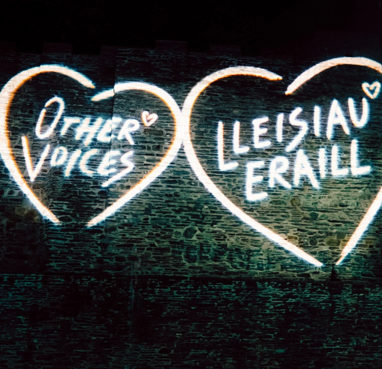 Projected images on a stone church wall - hearts with 'Other voices' and 'Lleisiau Eraill' text within.