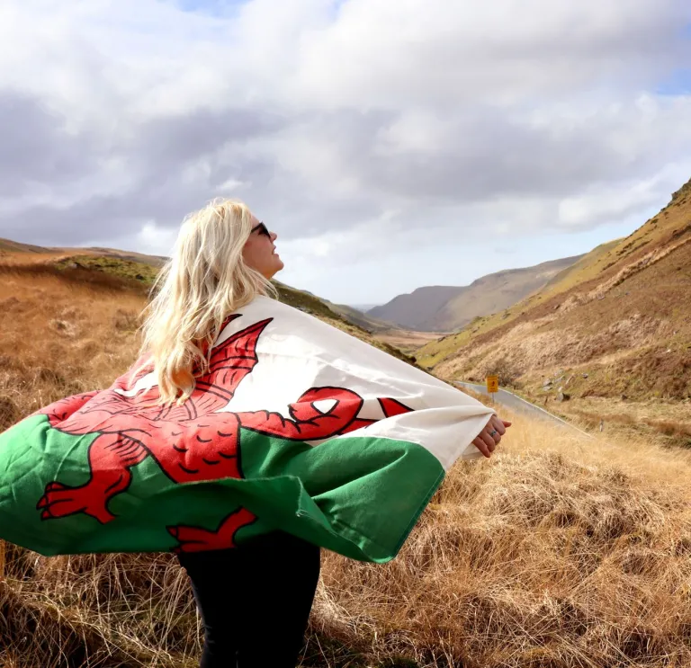 A person holding a Welsh flag over their shoulders looking out in awe over a grand mountainous landscape