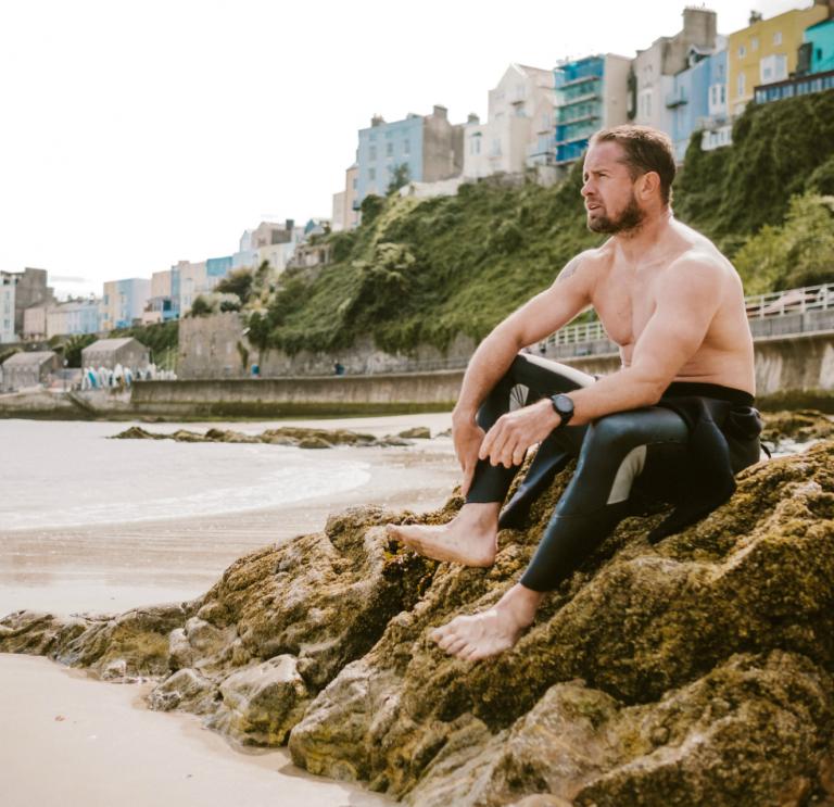 Shane Williams sentado en una roca en la playa del norte de Tenby con casas multicolores detrás.
