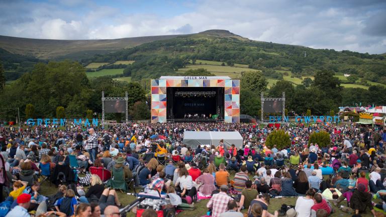 Eine Menschenmenge sitzt auf einem Feld und blickt auf eine Festivalbühne im Freien mit einem Hügel im Hintergrund