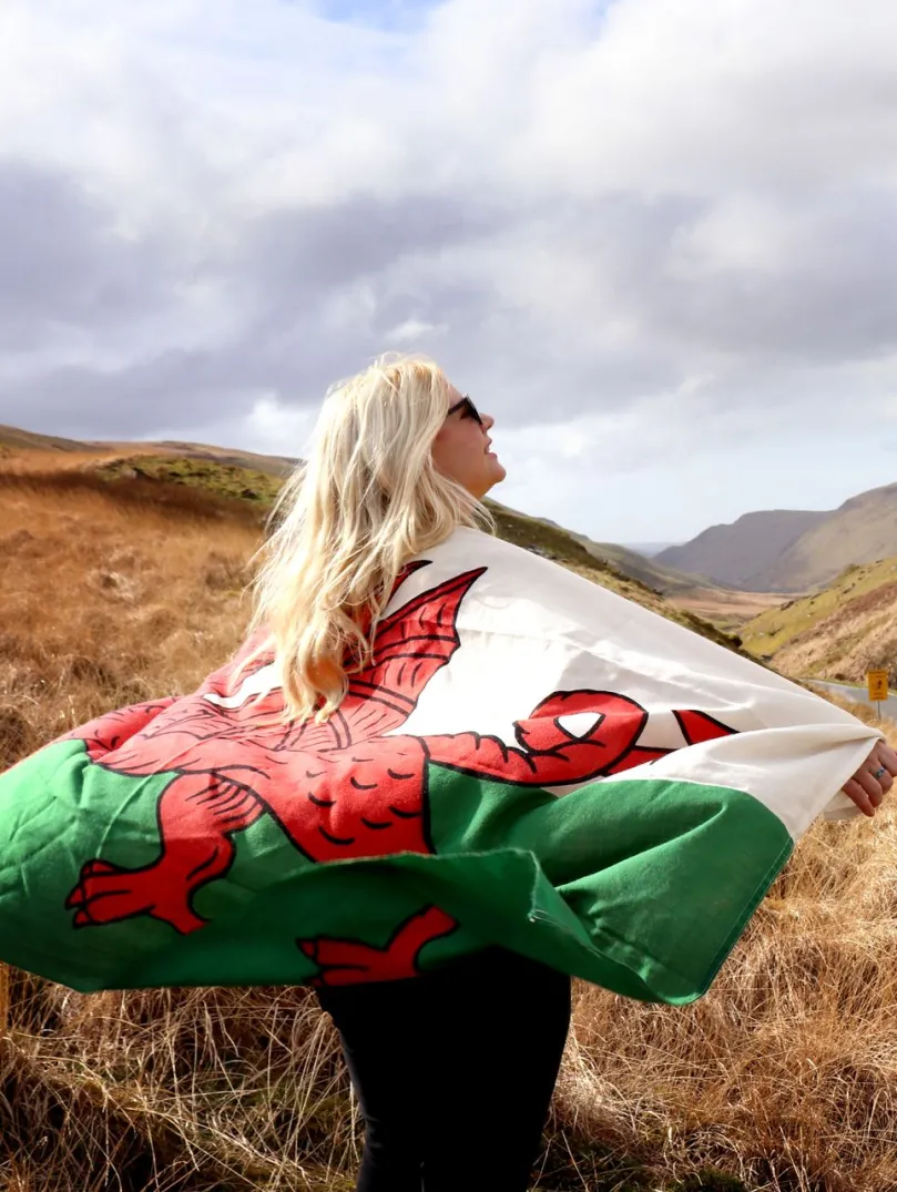 A person holding a Welsh flag over their shoulders looking out in awe over a grand mountainous landscape