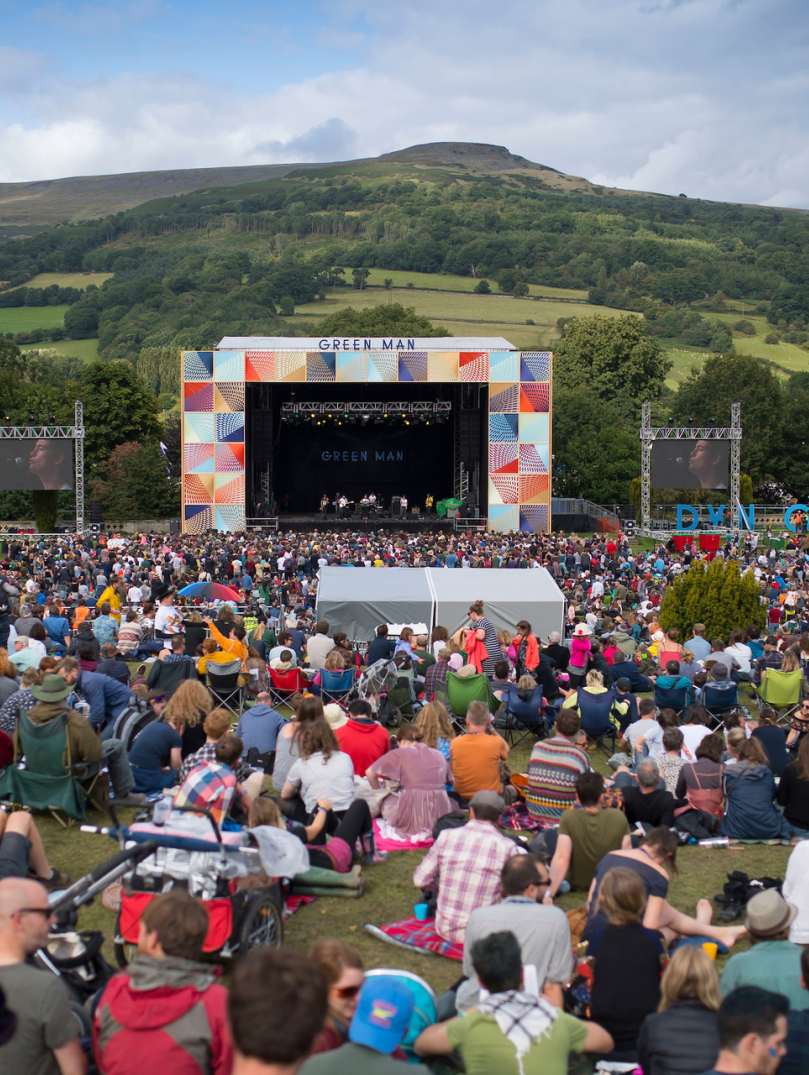 Eine Menschenmenge sitzt auf einem Feld und blickt auf eine Festivalbühne im Freien mit einem Hügel im Hintergrund