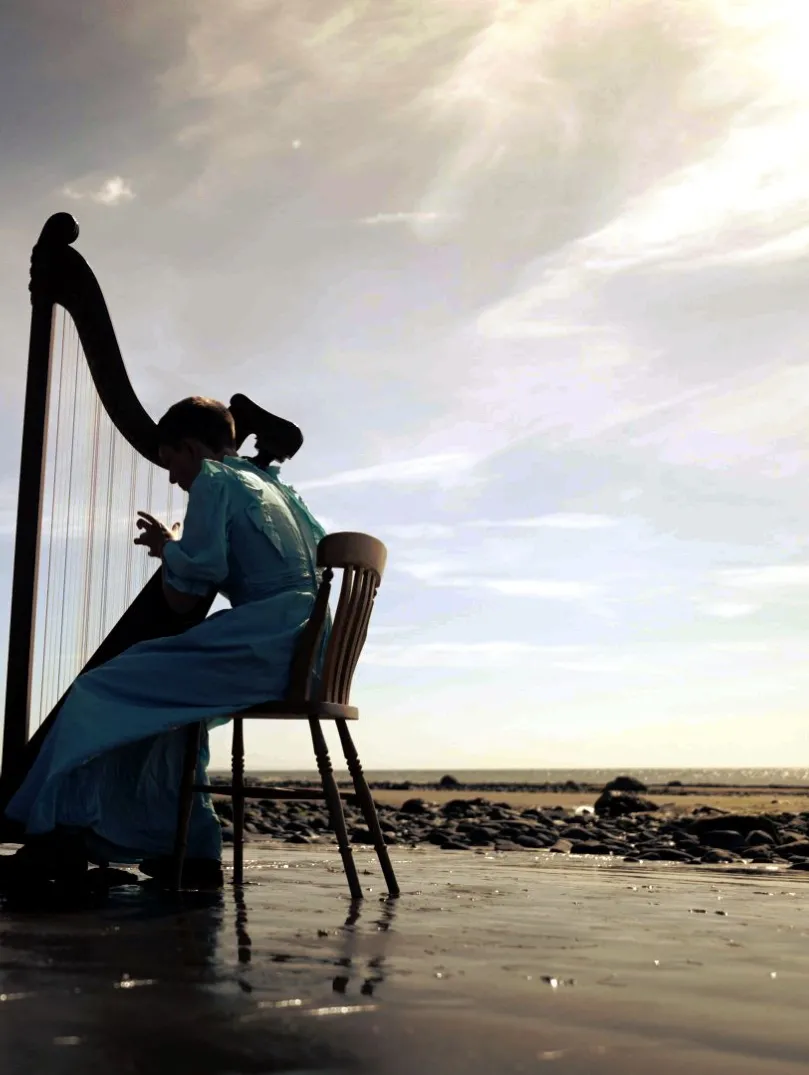 A young harp player wearing a long dress, sitting and playing a harp on a beautiful deserted beach