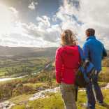 female and male walkers in the foreground looking down on a green valley  with a river