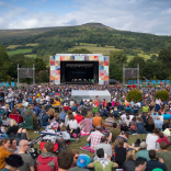 Eine Menschenmenge sitzt auf einem Feld und blickt auf eine Festivalbühne im Freien mit einem Hügel im Hintergrund