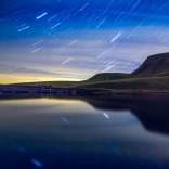 A night shot of a lake and some mountains with starry skies behind